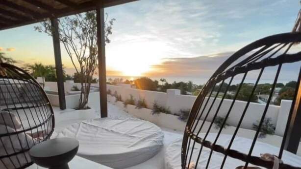 Eine Dachterrasse mit Blick auf den Sonnenuntergang, eine weiße Sitzgelegenheit mit Kissen und ein schwarzer Metallstuhl.