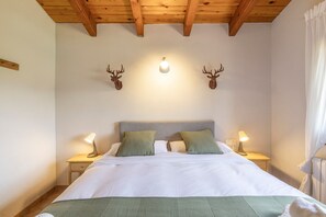 A bedroom with a wooden ceiling, a bed with white bedding, two bedside lamps, and two wall-mounted deer antlers.