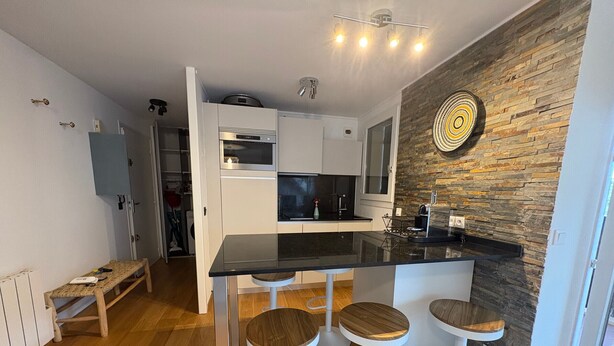 Una cocina moderna con encimera negra, armarios blancos y una pared de piedra. Hay tres taburetes y un banco.
