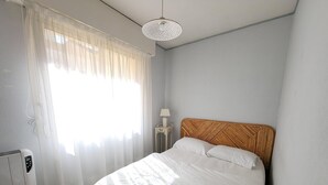 Un dormitorio con cama, una mesita de noche con lámpara, una ventana con cortinas translúcidas y una lámpara de techo.