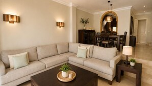 Una sala de estar con un sofá modular, una mesa de centro de madera oscura, dos lámparas de pared y un comedor con candelabro.