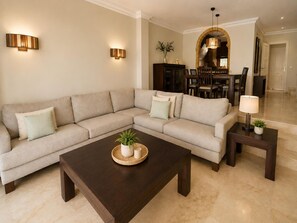 A living room with a sectional sofa, a dark wooden coffee table, two wall-mounted lamps, and a dining area with a chandelier. - House near the calm family beach (Estepona)