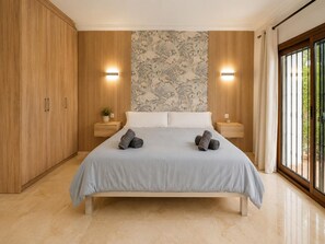 A modern bedroom with a large bed, wooden paneling, a floral wallpaper, two bedside lamps, and a view through a glass door. - House near the calm family beach (Estepona)