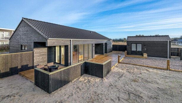 Una casa moderna frente a la playa con un amplio patio, un garaje y una edificación pequeña contigua.