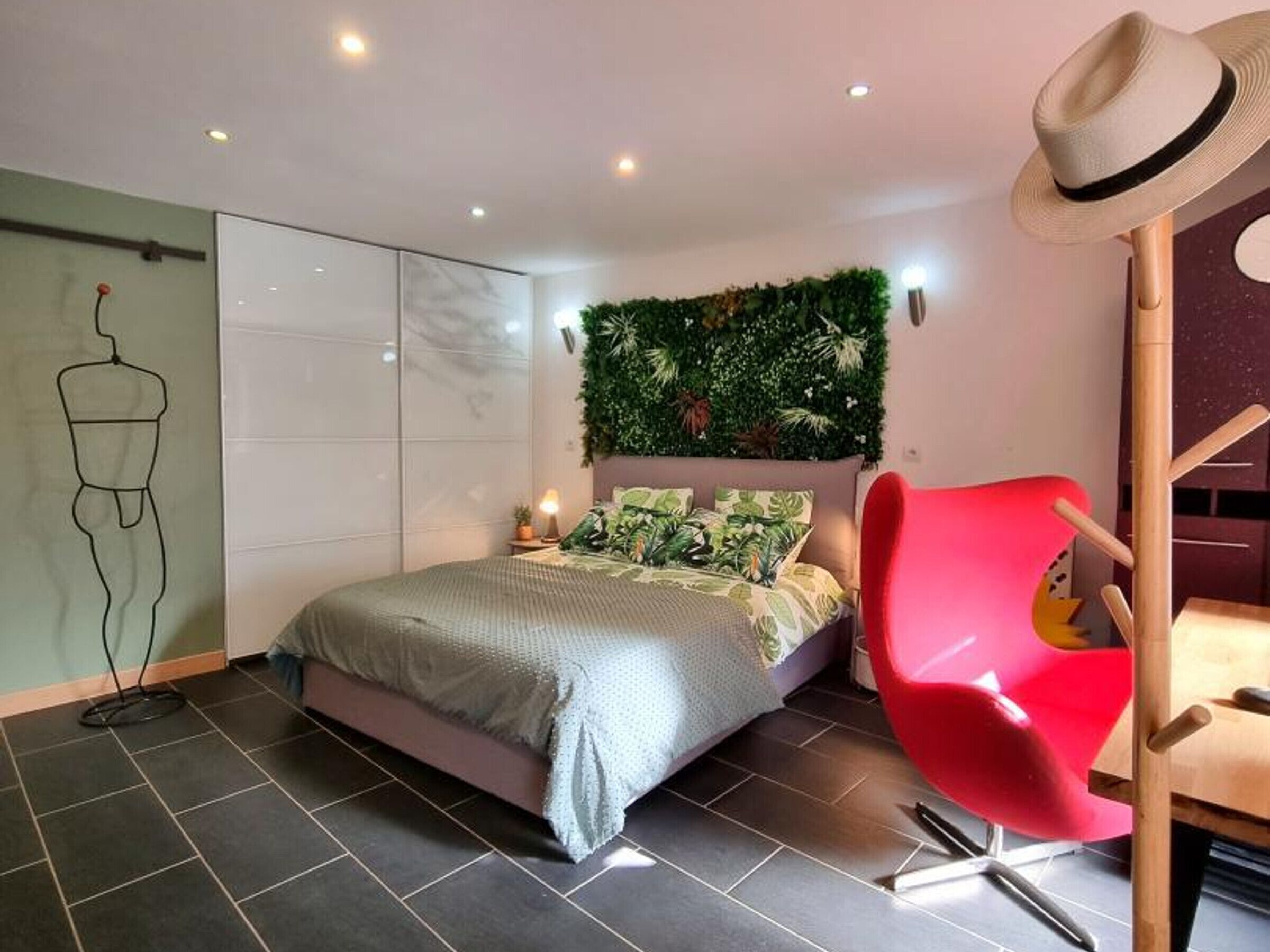 Une chambre moderne avec un lit, un fauteuil rouge, un chapeau posé sur un porte-chapeau et un mur orné d’une plante verte.