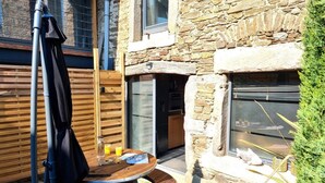 Een stenen huis met een houten terras, een ronde tafel met een stoel en een raam met uitzicht.