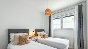 A bedroom with two beds, a nightstand with a lamp, a window with curtains, and a hanging light fixture. - Bright apartment, sea view - 4 guests (Estepona)