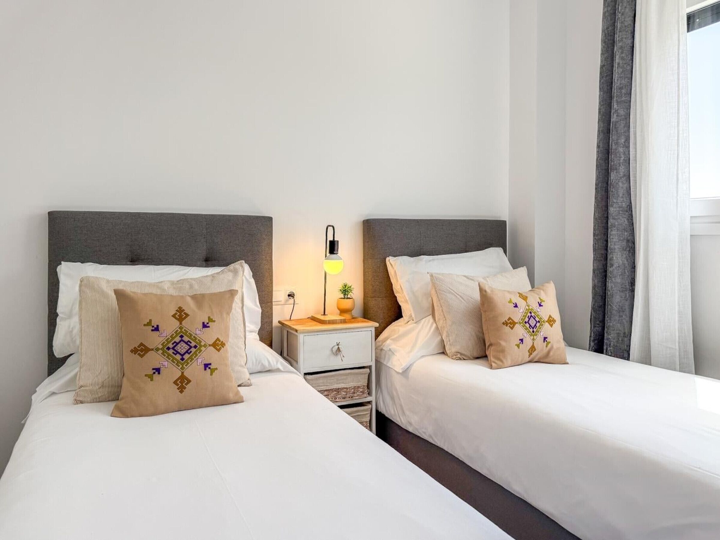 Habitación de hotel con dos camas, una mesita de noche con lámpara y una ventana con cortinas.