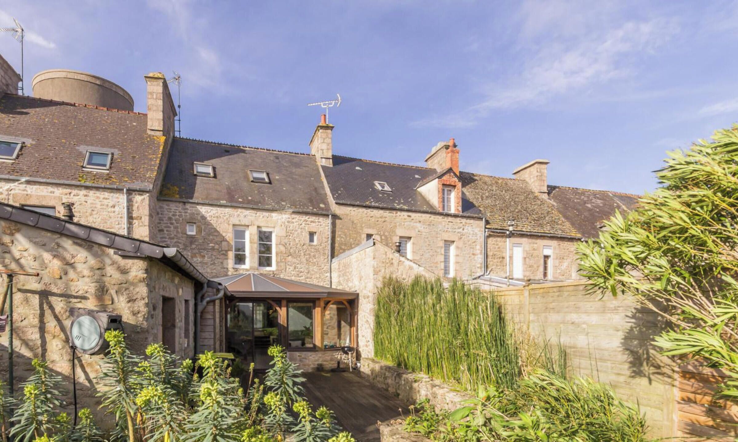 A stone building with multiple chimneys, a small attached structure with a roof, and a garden area with various plants.