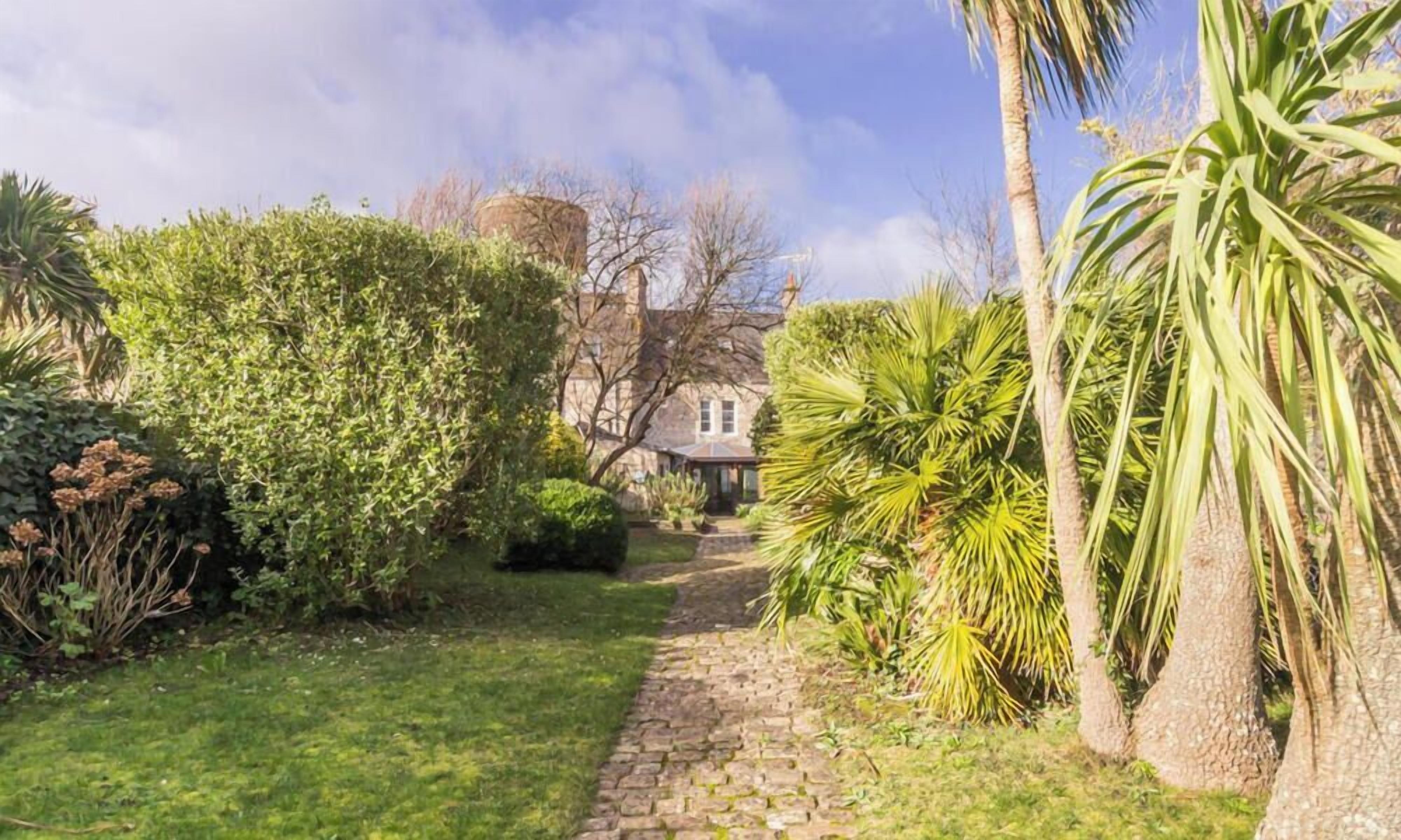 A garden pathway leading to a building with a stone tower, surrounded by lush greenery and palm trees.