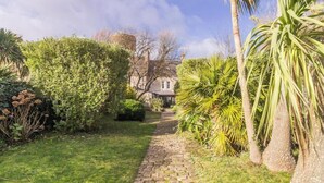A garden pathway leading to a building with a stone tower, surrounded by lush greenery and palm trees.