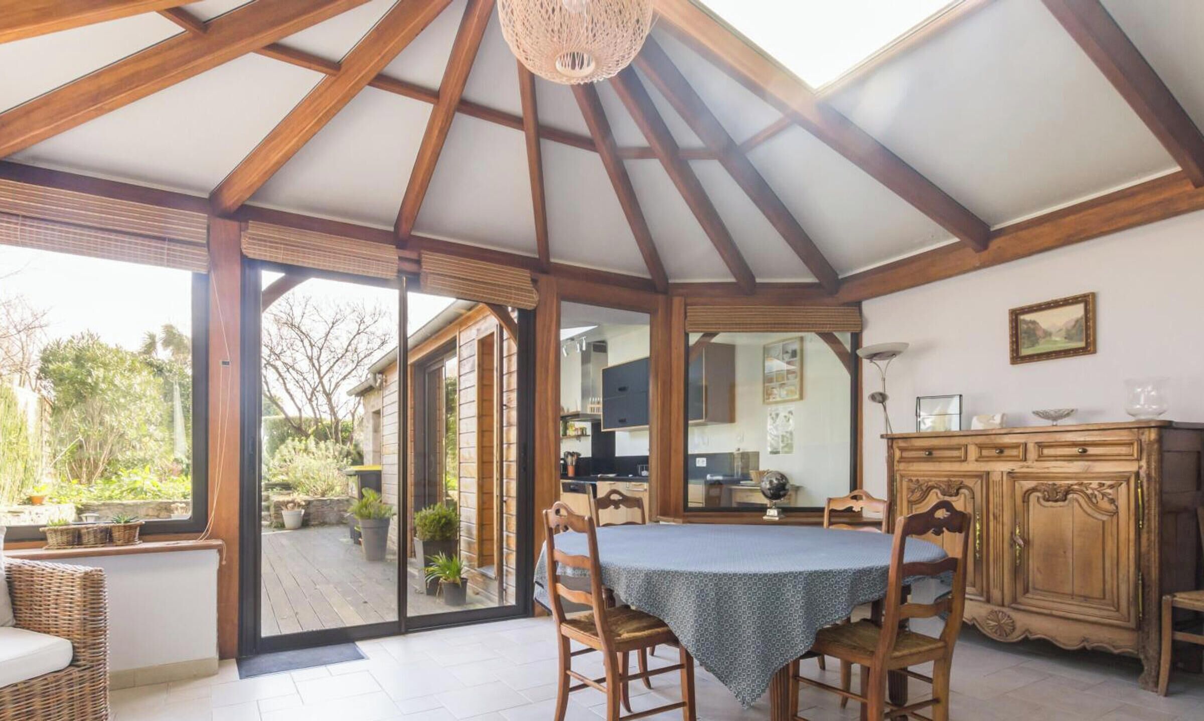 Une pièce spacieuse avec un plafond haut, des poutres en bois, une table à manger avec des chaises, un coin cuisine et une vue sur l’extérieur.