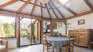 A spacious room with a high ceiling, wooden beams, a dining table with chairs, a kitchen area, and a view of the outdoors.