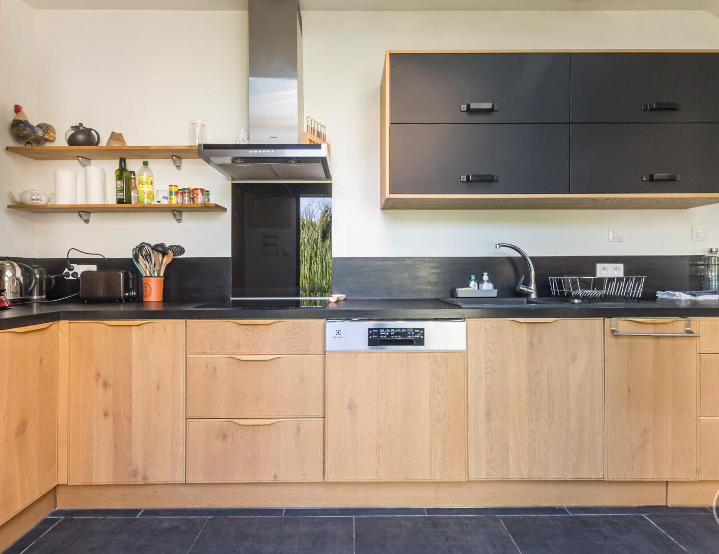 A modern kitchen with wooden cabinets, a black countertop, a built-in dishwasher, and a stainless steel range hood.
