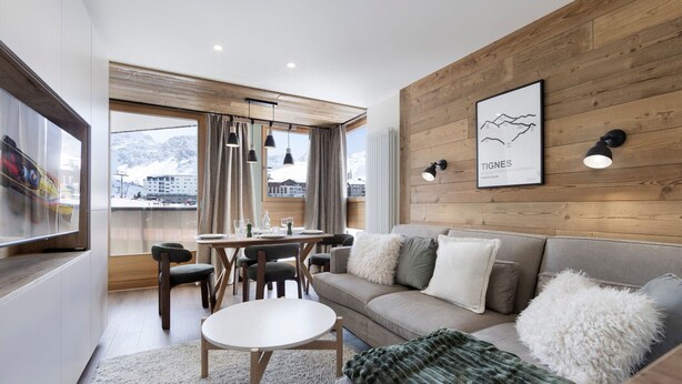 En moderne stue med trævæg, sofa, spisebord og udsigt til bjerge.