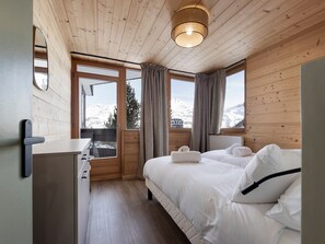 Ein Schlafzimmer einer Holzhütte mit einem Bett, einem Schminktisch, einem Spiegel und einem Fenster mit Blick auf schneebedeckte Berge.