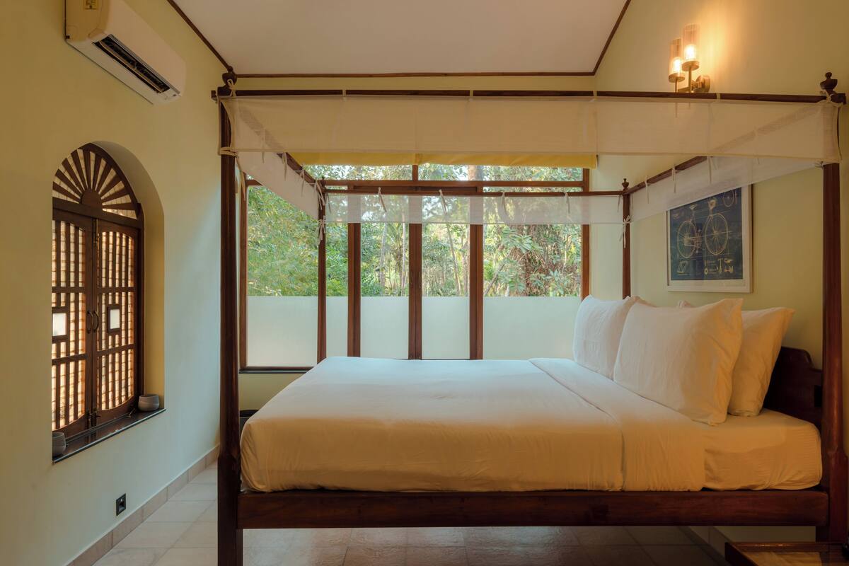 Quarto com cama, janela com vista para vegetação, ar condicionado e luminária na parede.