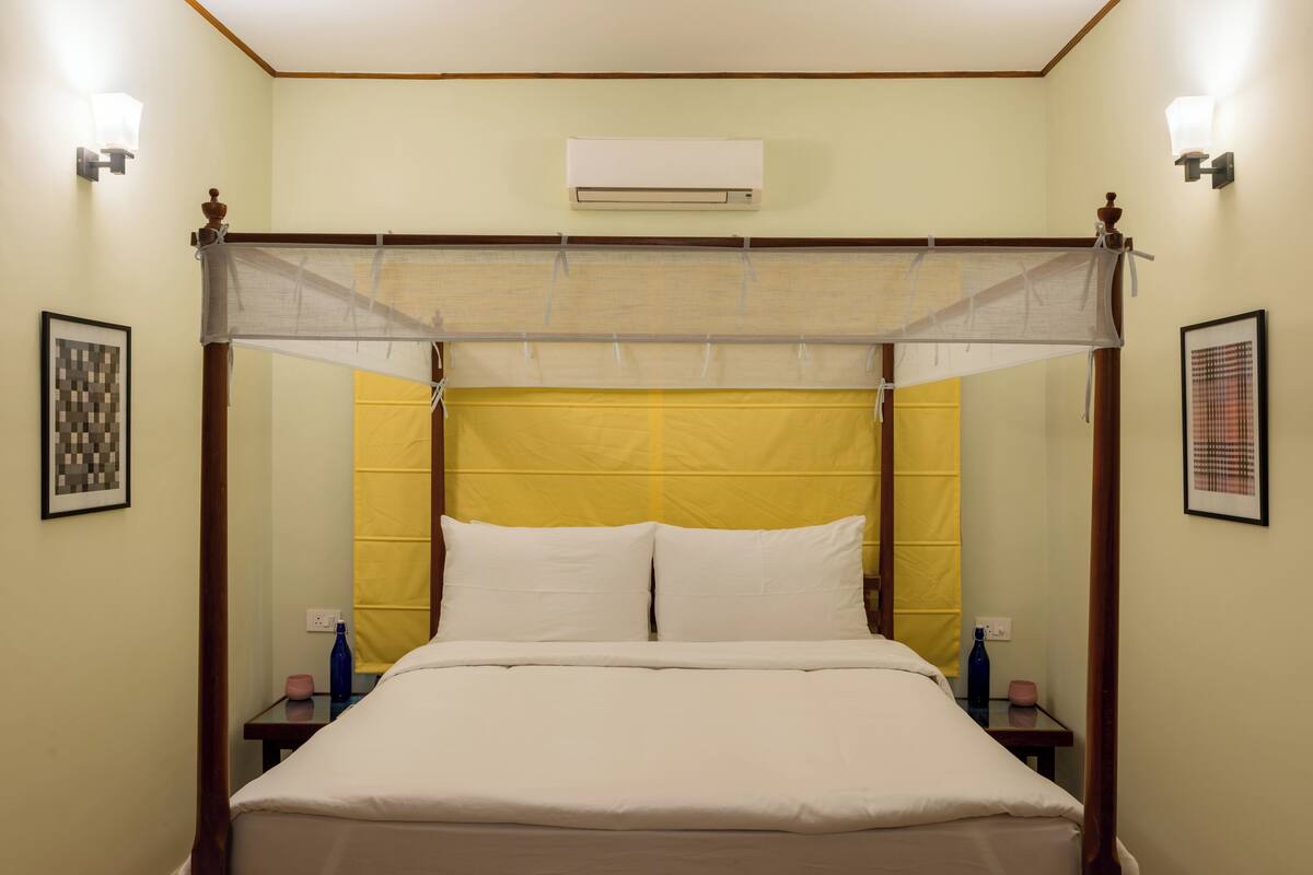 Uma cama com dossel e cabeceira amarela, duas mesas de cabeceira com garrafas e quadros nas paredes.