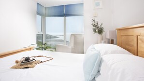 A modern hotel room with a large window, a bed, a chair, a bedside table with a lamp, and a view of the cityscape. - 18 The Sands - Sea View, Parking, Family friendly (Carbis Bay)