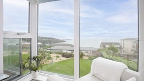 A room with large windows, a white sofa, and a view of a green landscape and buildings. - 18 The Sands - Sea View, Parking, Family friendly (Carbis Bay)