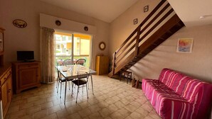 Un espace repas comprenant une table et des chaises, un canapé, un escalier et un téléviseur posé sur un meuble.