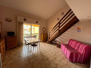 A dining area with a table and chairs, a sofa, a staircase, and a television on a cabinet. - Apartment with private parking - 4 guests (Fleury-Saint-Pierre-la-Mer)