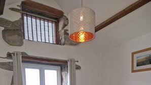 Une chambre à coucher comprenant un lit, une table de chevet, une lampe, une fenêtre avec des rideaux, un mur en pierre et un luminaire suspendu.