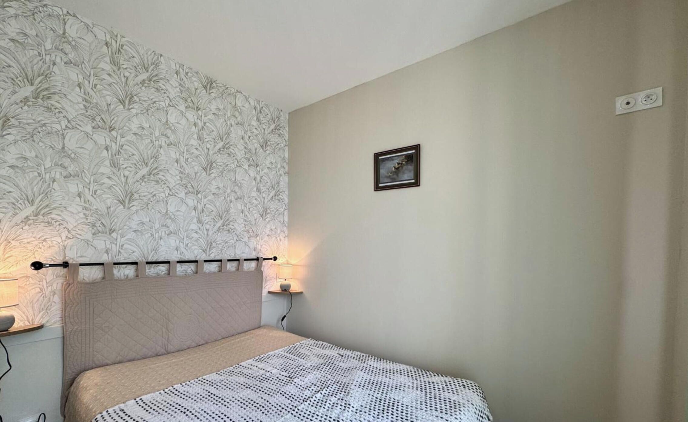 A bedroom with a patterned wallpaper, a bed with a knitted blanket, a bedside table with a lamp, and a small picture on the wall.