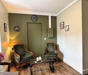 Une pièce comprenant deux fauteuils verts, une table basse en bois avec des bougies, une horloge murale et des tableaux encadrés.