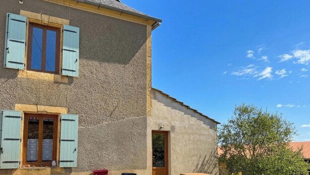 Un edificio de dos pisos con contraventanas azul claro, fachada de piedra y un cielo azul despejado.