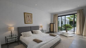 Un dormitorio moderno con una cama grande, mesitas de noche, un cuadro y vistas al exterior con zonas verdes.