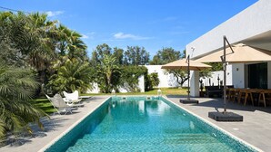 Piscina con sillas de descanso y sombrillas, rodeada de vegetación y un edificio blanco.