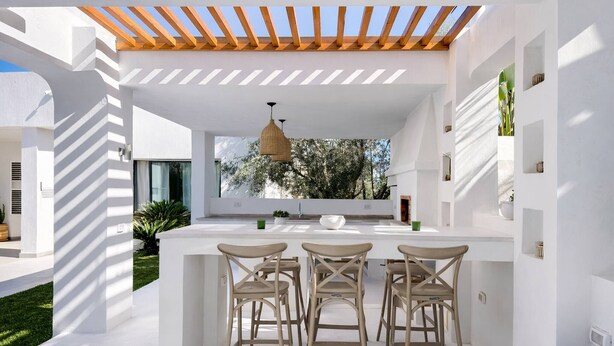 Una cocina moderna con una isla central, taburetes de bar, iluminación colgante y una pérgola.