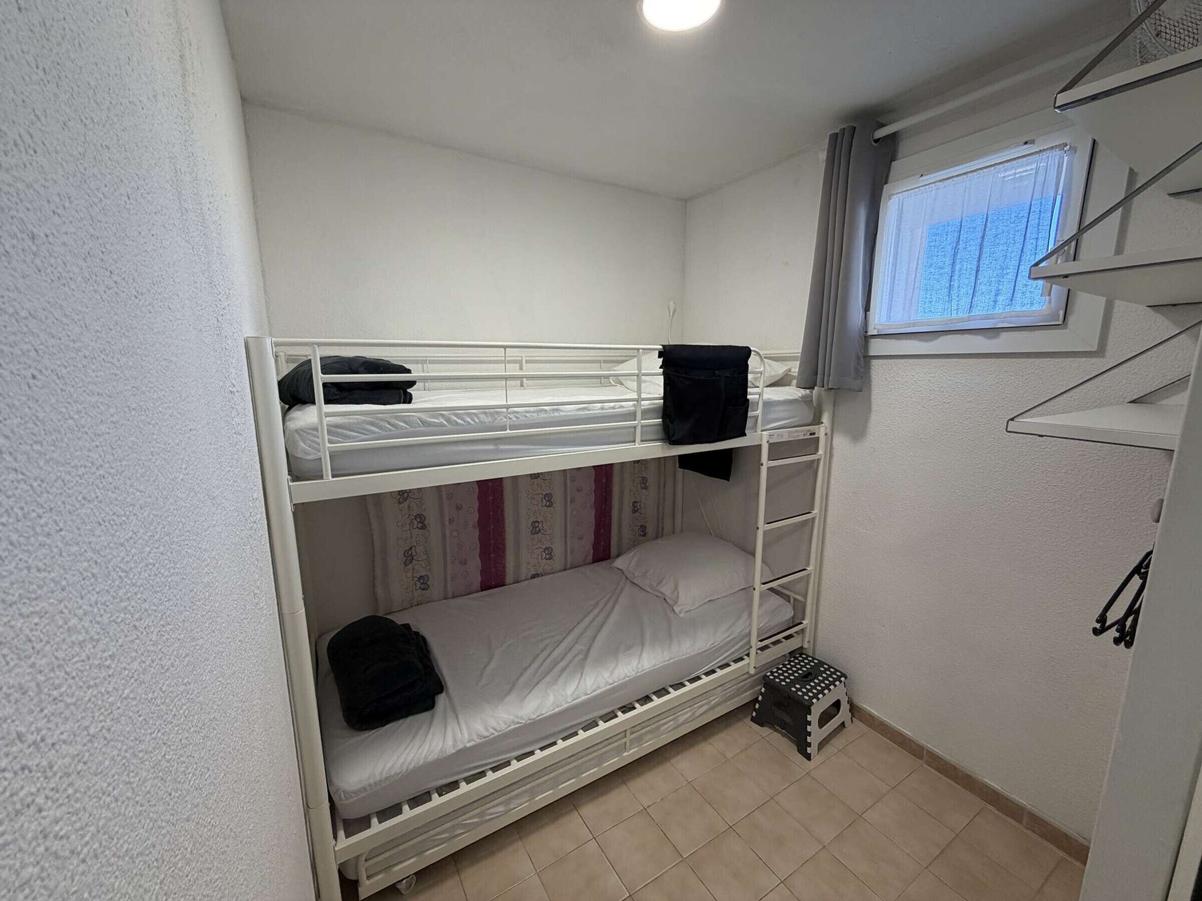 Dormitorio con literas, una ventana, un taburete pequeño y una estantería.