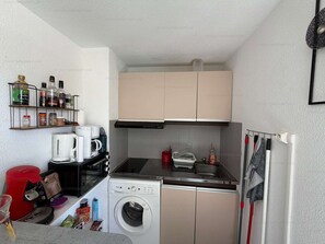 A compact kitchen with a washing machine, coffee maker, and a small shelf with various items. - Cozy apartment with balcony - 4 guests (Narbonne)
