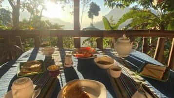 Mesa al aire libre con diversos platos, una tetera y tazas, rodeada de exuberante vegetación y montañas al fondo.