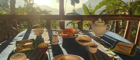 A table set outdoors with various dishes, a teapot, and cups, surrounded by lush greenery and mountains in the background.