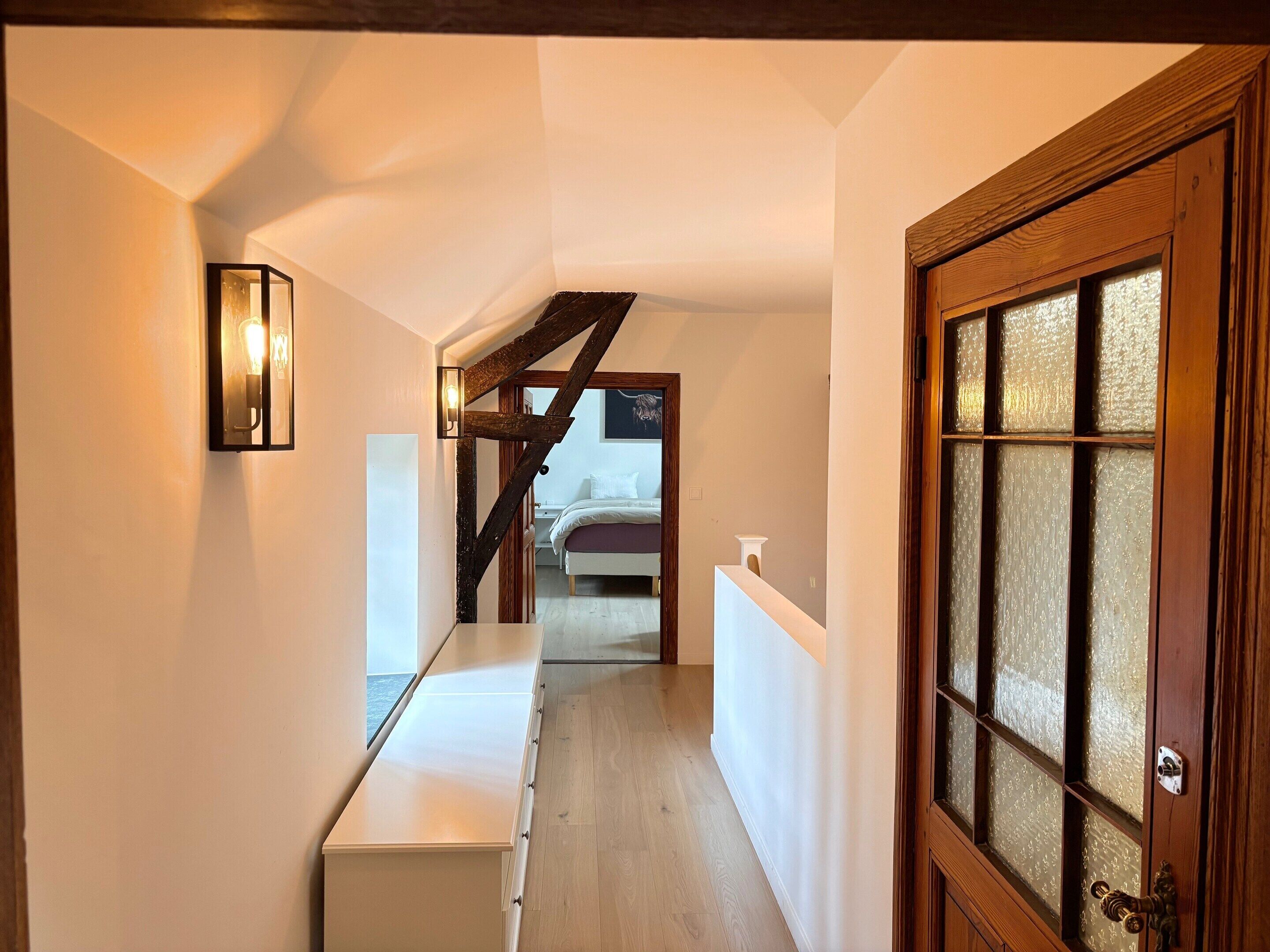 Ein schmaler Flur mit einer Holztreppe, einer weißen Tür mit mattiertem Glas und einem Bett im Hintergrund.