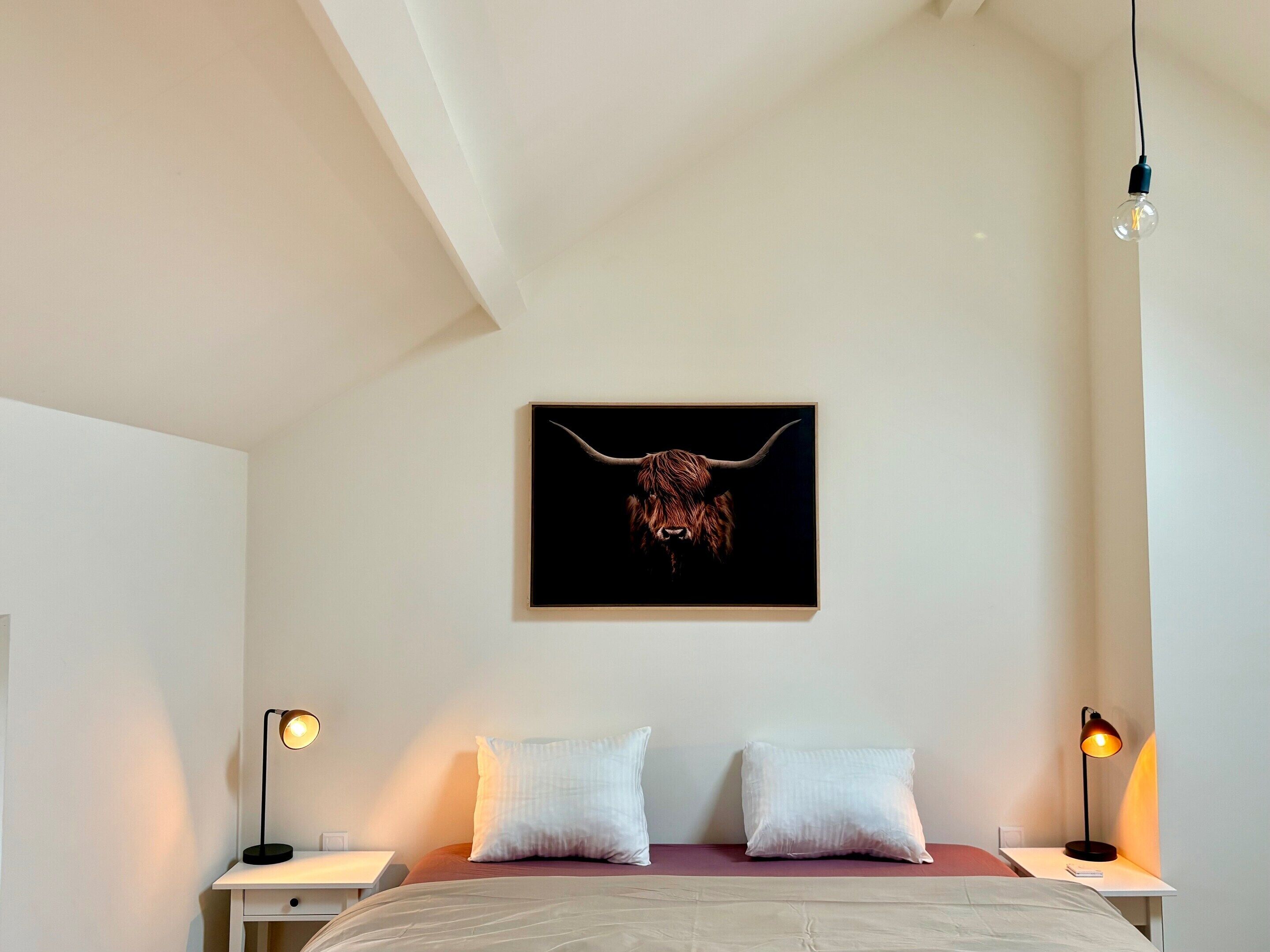Ein Schlafzimmer mit einem Bett, zwei Nachttischlampen, einem Wandbild mit einem Stierkopf und einer weißen Decke.