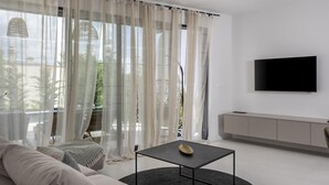 Ein modernes Wohnzimmer mit einer Couch, einem Couchtisch, einem an der Wand befestigten Fernseher und Blick durch transparente Vorhänge.