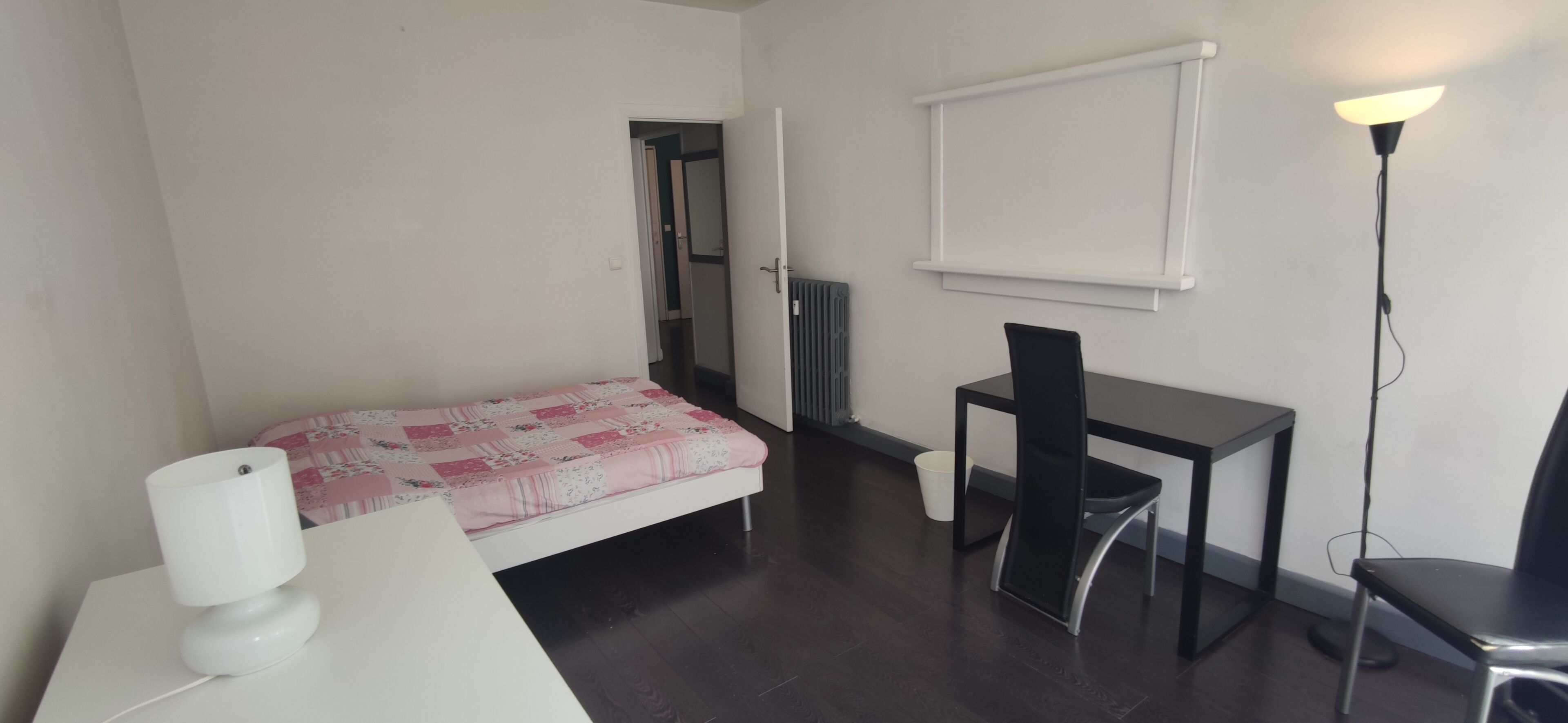 Een kleine, eenvoudig ingerichte kamer met een bed, een bureau, een stoel, een radiator en een vloerlamp.