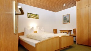 A wooden bed with white bedding, a small table, a chair, and a wardrobe in a room with white walls and a wooden ceiling.