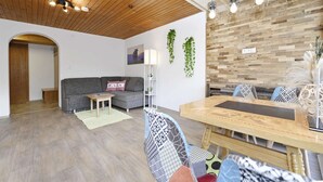 A modern living space with a wooden ceiling, a grey sofa, a dining table with patterned chairs, and a wall-mounted light fixture.