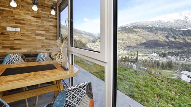 A modern interior with a wooden wall, a dining area with a table and chairs, and a large window offering a view of a mountainous landscape.