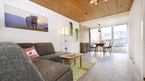 A modern living room with a sofa, a dining table, and chairs. There is a large window with a view of mountains.