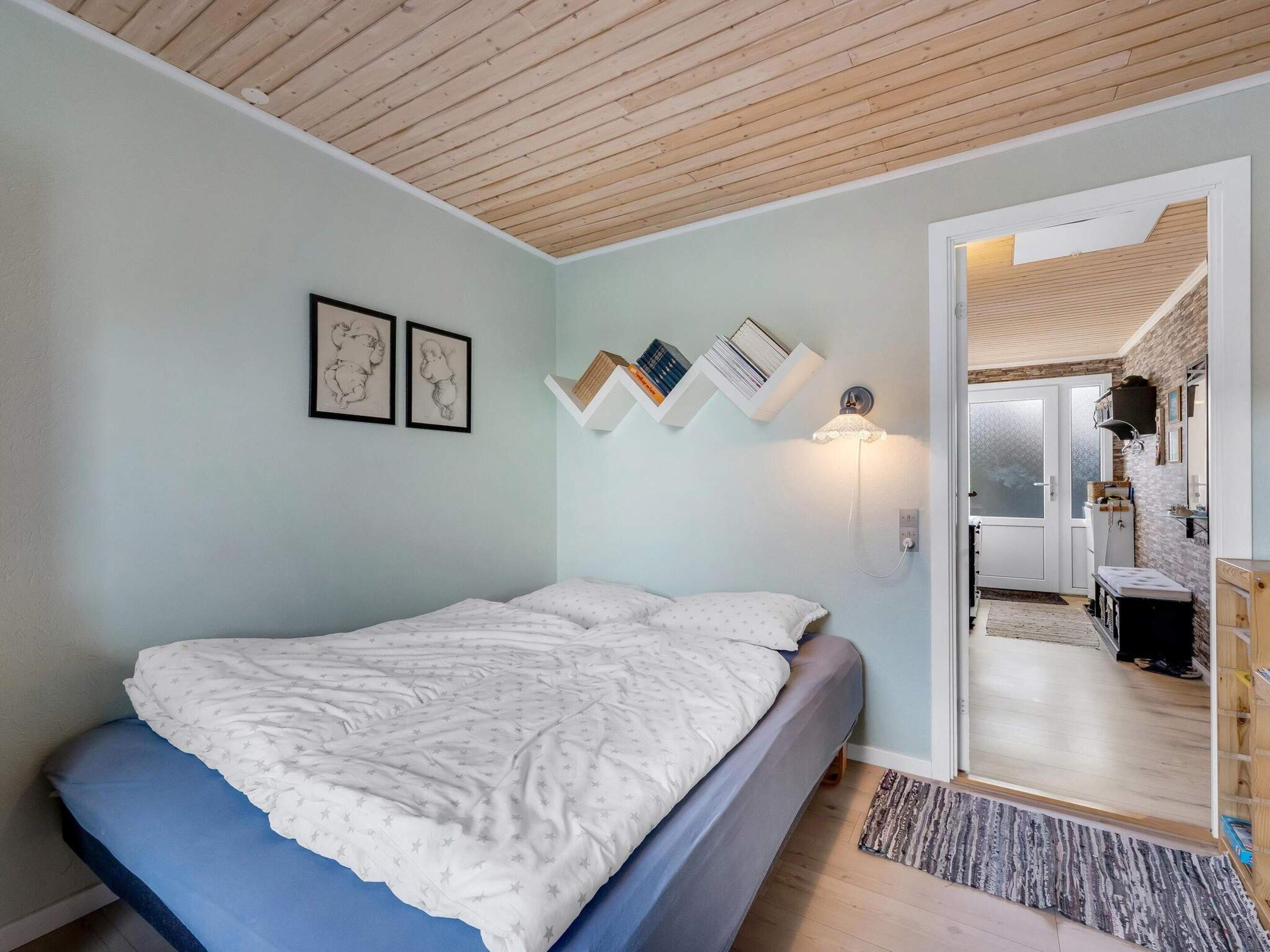 A bedroom with a bed, a wall-mounted shelf with books, a wall-mounted light, and a door leading to a bathroom.