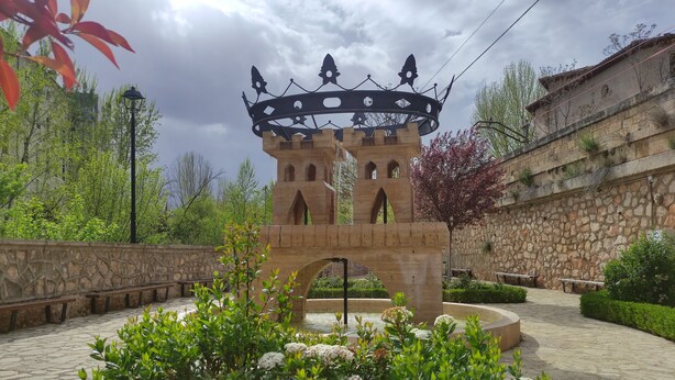 Un puente de piedra con un arco decorativo, rodeado de vegetación y un muro de piedra.