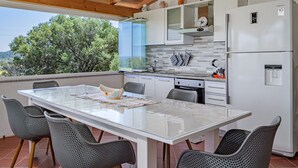 Une cuisine avec un réfrigérateur blanc, un four à micro-ondes et un évier. On y trouve une table avec des chaises et une vue sur les arbres à l’extérieur.