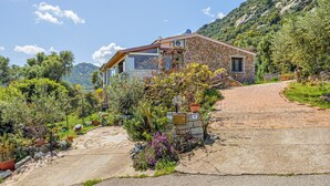Une maison en pierre avec une allée pavée, entourée d’une végétation luxuriante et adossée à une montagne.