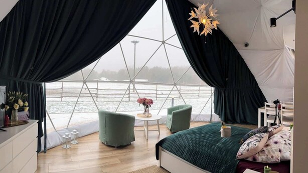 A tented room with a bed, two chairs, a small table, and a view of a body of water.